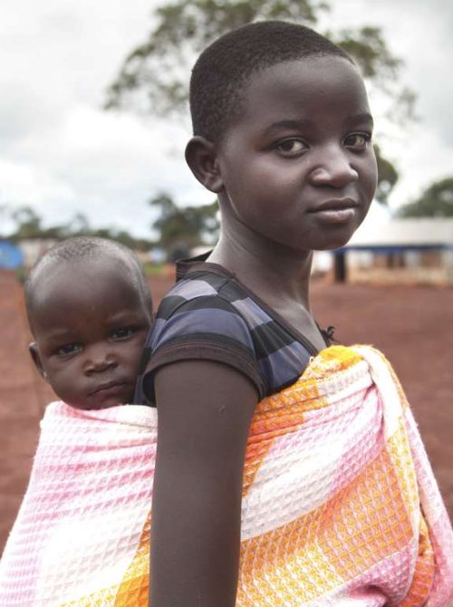 A young girl carries a baby on her back at the Save the Children-supported Nyarugusu Camp for refugees from Burundi. Save the Children created child-friendly spaces at the camp to help children play, express themselves and recover from their traumatic experiences. Photo credit: Martin Kharumwa/Save the Children, April 2016.